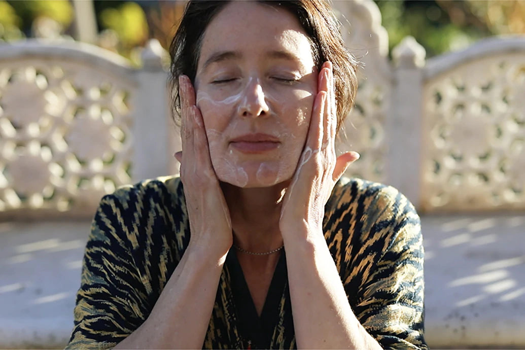 Woman gently massaging cleanser into her face while seated outdoors on a patterned stone bench in soft sunlight