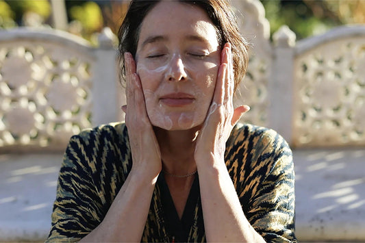Woman gently massaging cleanser into her face while seated outdoors on a patterned stone bench in soft sunlight