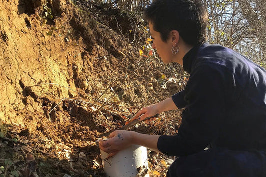 Person crouching outdoors scraping clay from a hillside into a white bucket