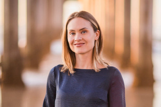 Portrait of a woman smiling in a navy top beneath a sunlit pier with warm blurred columns in the background