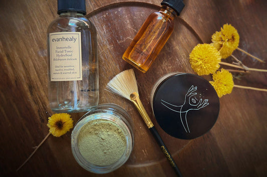 Flat lay of evanhealy skincare: Immortelle facial tonic, powder mask, amber facial oil, brush, and yellow flowers on wood