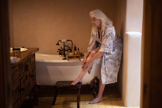 Woman in a patterned robe massaging oil into her lower leg beside a freestanding tub in a warm, softly lit bathroom