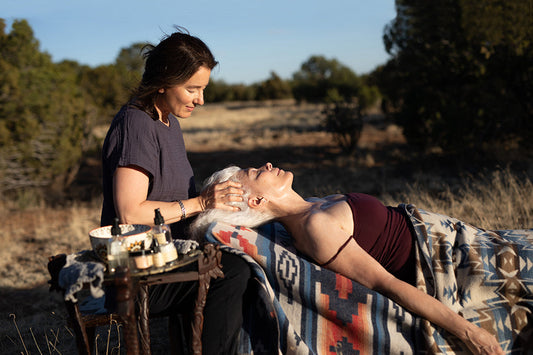 Esthetician giving a facial massage to an older woman lying on a patterned blanket outdoors in golden light