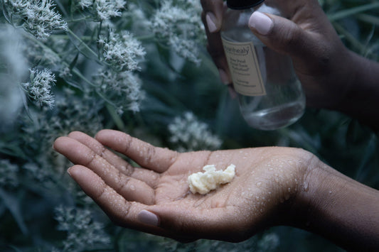 Close-up of wet hands holding shea butter with a clear skincare bottle above, dew-covered white flowers in background