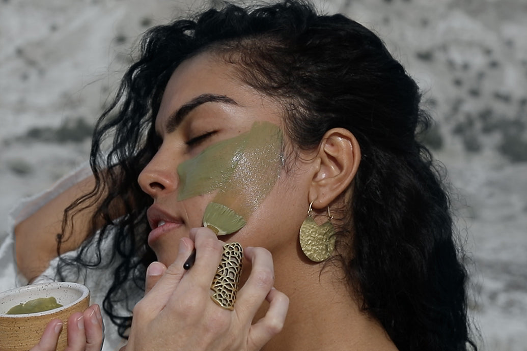 Woman having green clay facial mask painted onto her cheek with a brush, natural outdoor background