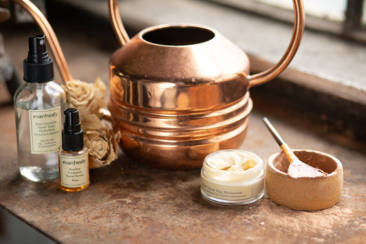 Evanhealy skincare bottles and an open moisturizer jar on a rustic windowsill with a copper watering can and dried roses