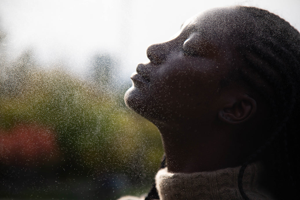 Side profile of a person tilting their face into a fine spray of water outdoors, backlit by soft light