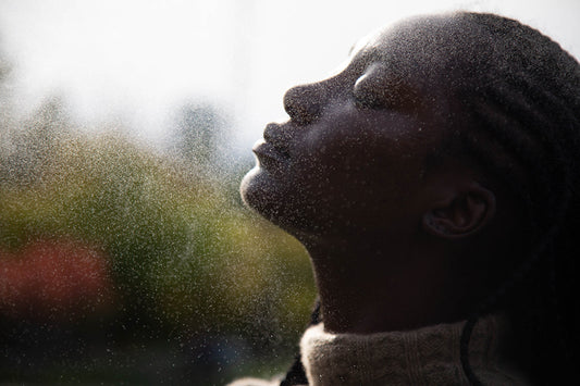 Side profile of a person tilting their face into a fine spray of water outdoors, backlit by soft light
