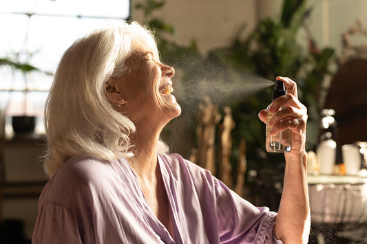 Mature woman smiling as she sprays Hydrosoul facial mist toward her face in a sunlit bathroom