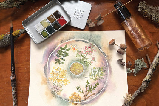 Watercolor floral wreath painting on paper with a brush, metal watercolor palette, glass roller bottle, and moss on a wooden table