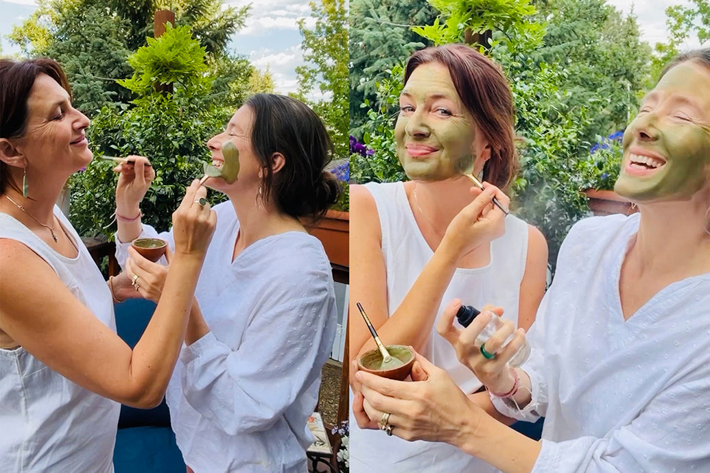 Two women applying green tea clay and aloe vera face masks together on a sunny outdoor patio, smiling