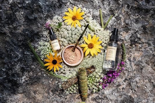 Evanhealy skincare bottles, a bowl of powdered mask with brush, and a sage bundle arranged with wildflowers on a rock, evoking a calming ritual
