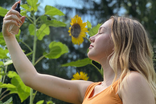 Young woman with eyes closed spraying a facial mist toward her face while standing in a sunflower garden