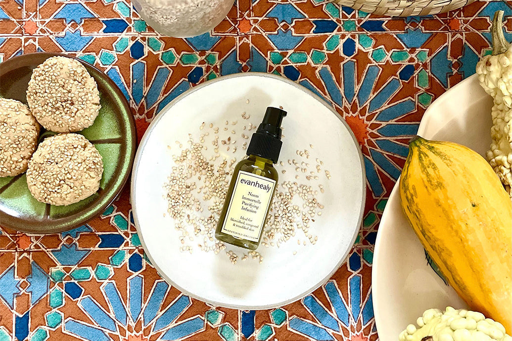 Top-down of evanhealy Neem Immortelle Purifying Infusion bottle with pump on white plate with sesame seeds on patterned tile