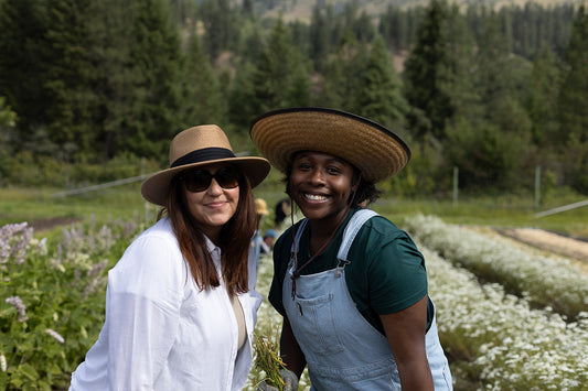Two farm workers wearing wide-brim hats smiling in a flower field with forested hills in the background