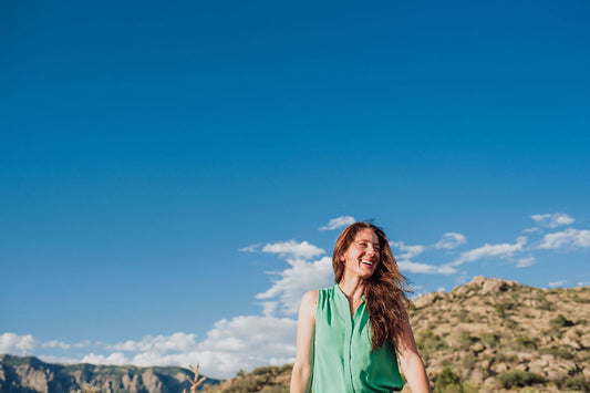 Woman smiling outdoors with windswept hair, standing in front of rocky hills under a clear blue sky