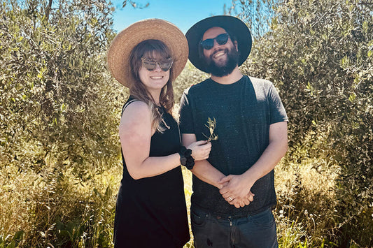 Two people smiling in an olive grove, woman in a straw hat holding an olive branch and man wearing a dark shirt and wide-brimmed hat