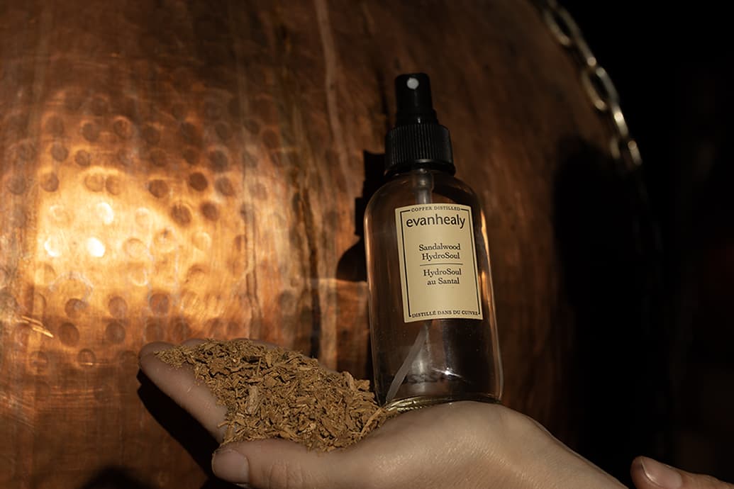 Front view of Sandalwood HydroSoul spray bottle with label and sandalwood shavings in hand against copper background