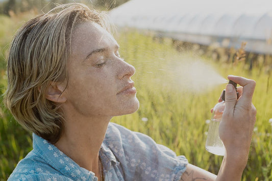 Woman spraying facial mist on her face in a sunlit grassy field