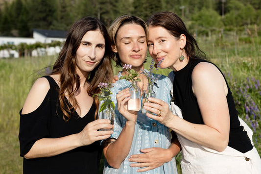 Three women in a sunlit meadow holding small glass bottles with fresh flowers and a light-pink HydroSoul serum, smiling and embracing