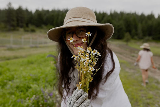 Woman holding chamomile flowers in a field, wearing a wide-brimmed hat and gardening gloves
