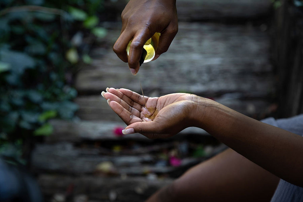 Hands pouring golden body oil into a cupped palm over a weathered wooden deck
