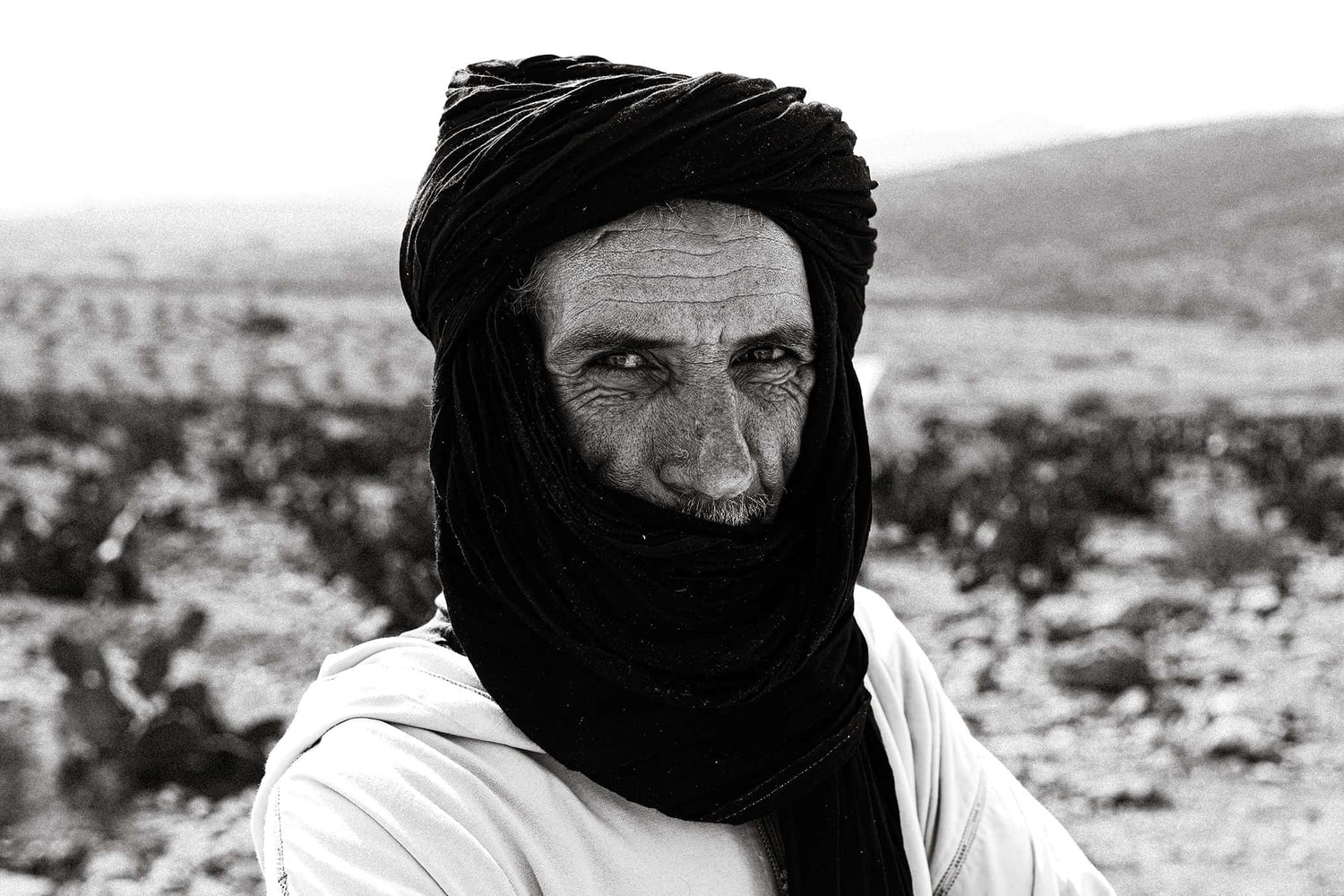 Man wearing a dark turban and white robe looking at the camera in a rocky Moroccan landscape