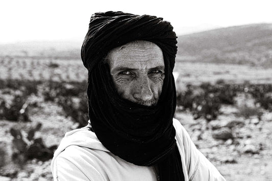 Man wearing a dark turban and white robe looking at the camera in a rocky Moroccan landscape