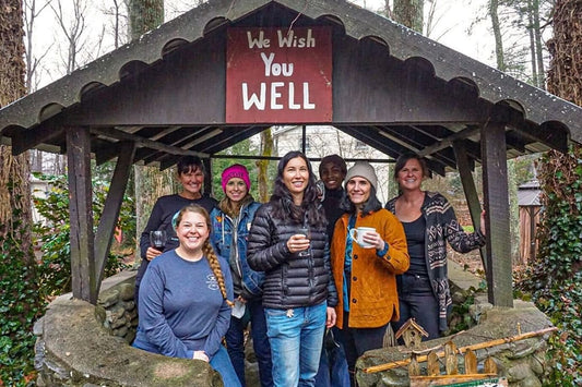 Seven people smiling and holding cups under a wooden shelter that reads ‘We Wish You Well’ in a wooded yard