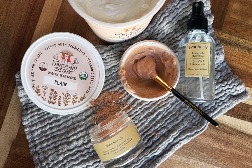 Bowl of French rose clay mixed with plain yogurt and a brush beside an evanhealy Immortelle HydroSoul spray and yogurt tub on a textured towel