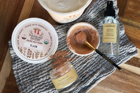 Bowl of French rose clay mixed with plain yogurt and a brush beside an evanhealy Immortelle HydroSoul spray and yogurt tub on a textured towel