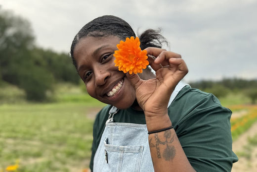 Smiling woman holding an orange calendula flower to her face in a field, wearing denim overalls and a green shirt