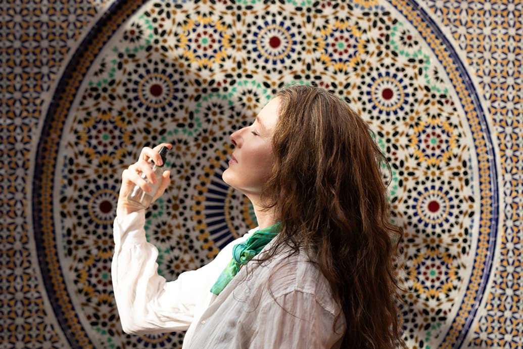 Woman spraying a facial mist toward her face in front of a decorative Moroccan tile mosaic