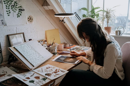 Robin Elise Pieterse painting at a wooden desk in a sunlit attic studio surrounded by sketchbooks, brushes, mugs, and potted plants