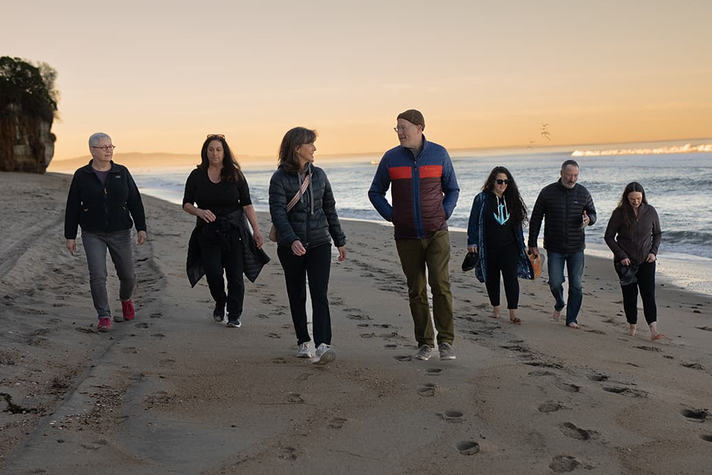 Seven people walking along a beach at sunset, talking and leaving footprints in the sand
