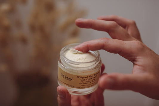 Close-up of open jar of evanhealy Whipped Shea Butter with Olive Leaf, fingertip scooping cream, blurred neutral background