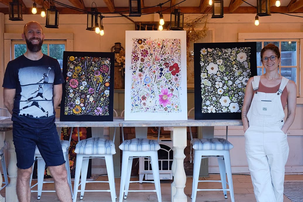 Keith and Rachel of Flower Press Studio standing at a table displaying three framed floral prints in their studio with hanging lights and stools