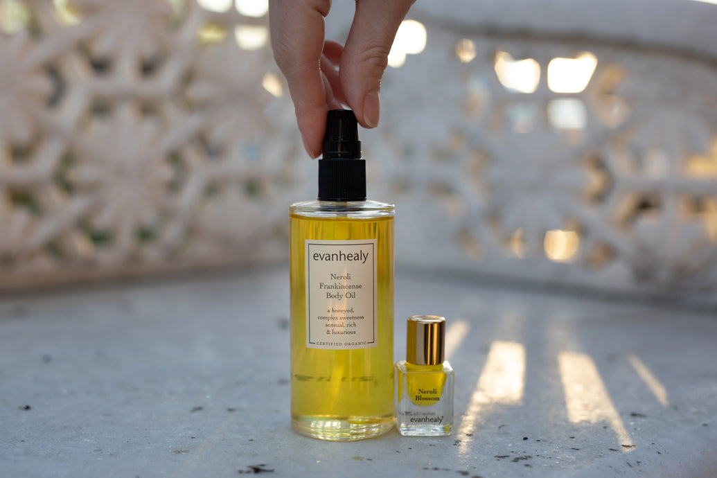 Front view of evanhealy Neroli Frankincense body oil bottle with pump and small Neroli Blossom perfume on sunlit carved stone