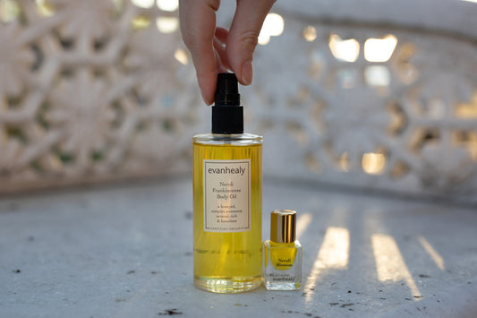 Front view of evanhealy Neroli Frankincense body oil bottle with pump and small Neroli Blossom perfume on sunlit carved stone