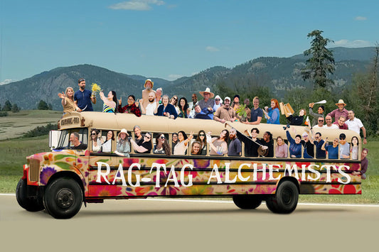 Tie-dye painted bus labeled RAG-TAG ALCHEMISTS filled with smiling people on the roof and inside, on a road with mountains