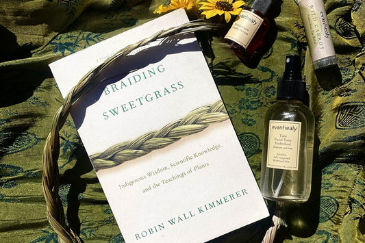 Book ‘Braiding Sweetgrass’ on green fabric with a braided grass crown and evanhealy facial tonic and small skincare jars in sunlight