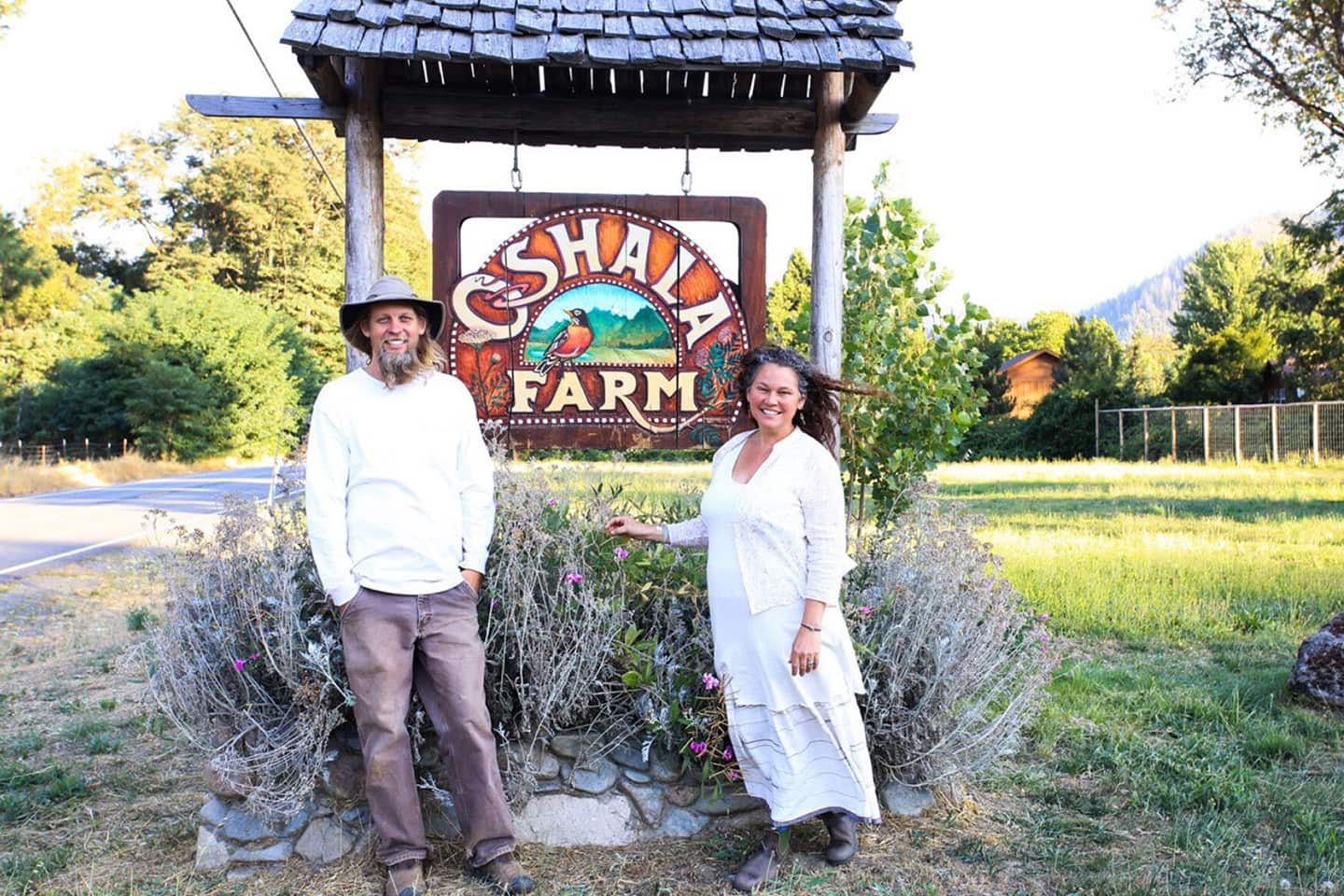 Two people smiling beside a wooden Oshala Farm sign at a farm entrance with lavender beds and trees in the background