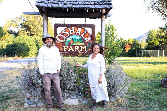 Two people smiling beside a wooden Oshala Farm sign at a farm entrance with lavender beds and trees in the background
