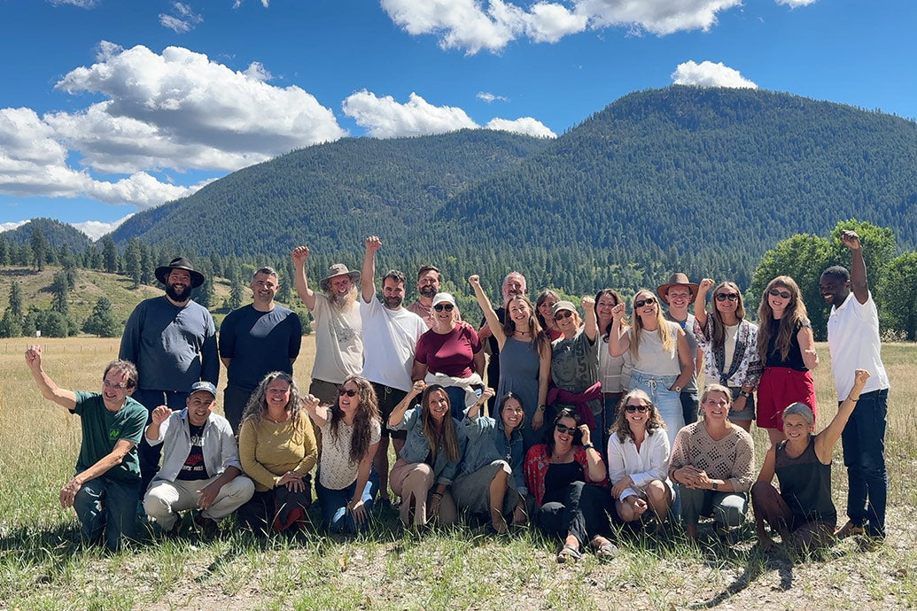 Group of about 25 people raising their fists and smiling in a sunny mountain meadow with forested hills behind them