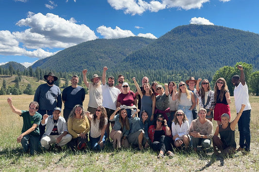 Group of about 25 people raising their fists and smiling in a sunny mountain meadow with forested hills behind them