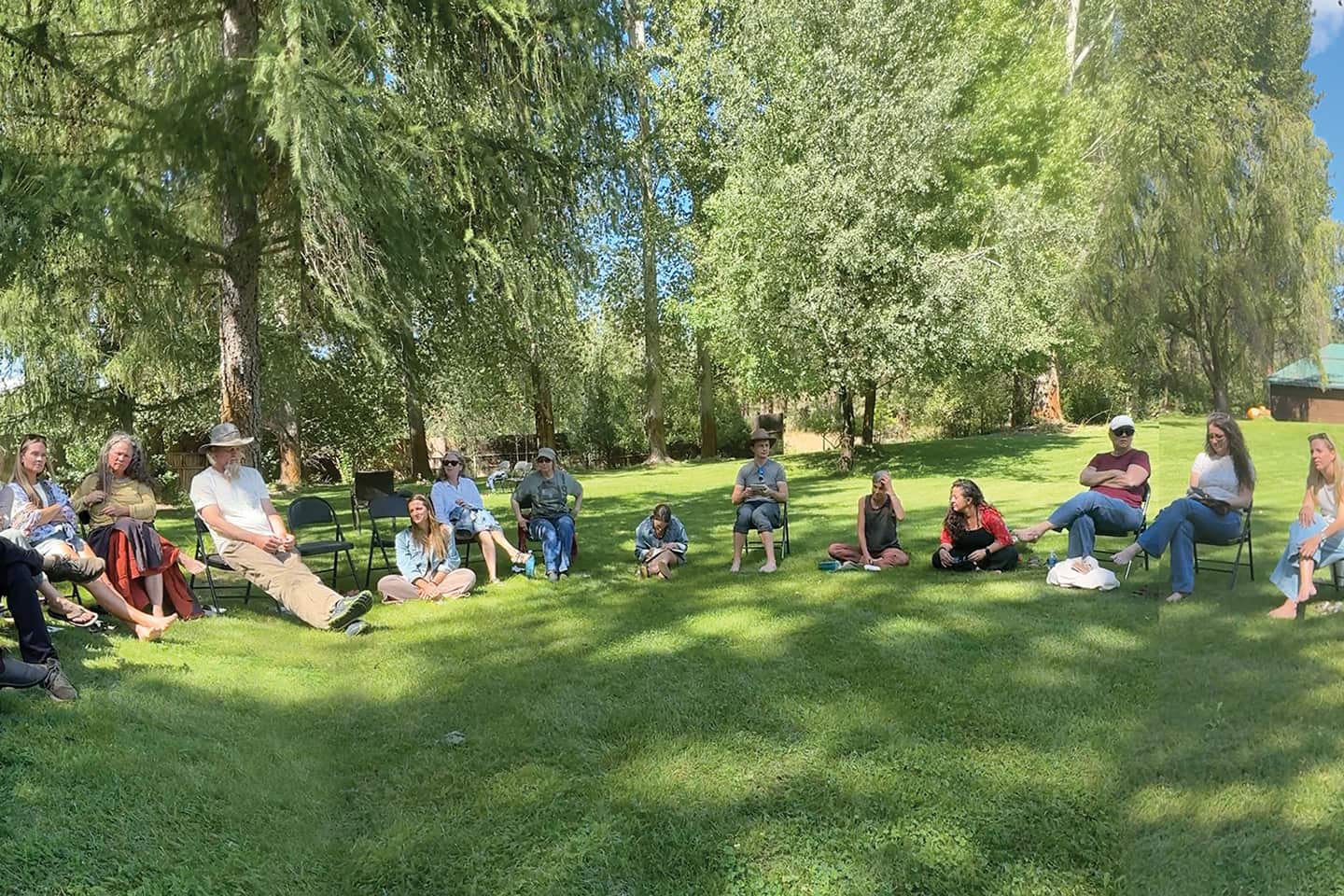 Group of people sitting in a wide circle on a grassy lawn under tall trees at a sunny outdoor gathering