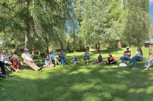 Group of people sitting in a wide circle on a grassy lawn under tall trees at a sunny outdoor gathering