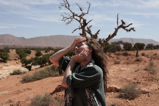 Woman shielding her eyes and touching her face in a sunlit Moroccan desert with dry trees and distant hills
