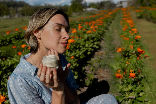 Woman with eyes closed holding a jar of face cream to her chin while sitting in a field of orange calendula flowers