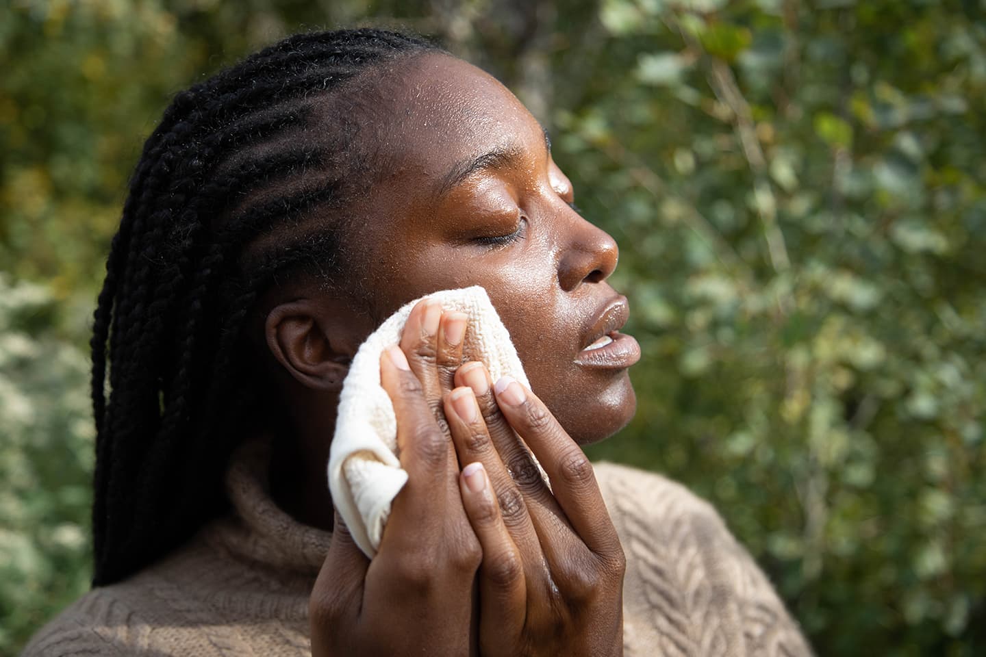 Close-up of a woman gently wiping oil from her face with a cotton cloth outdoors against a leafy green background